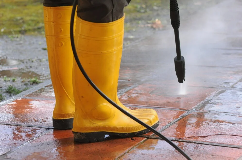 Home cleaning involves pressure washing a surface while wearing yellow rubber boots.